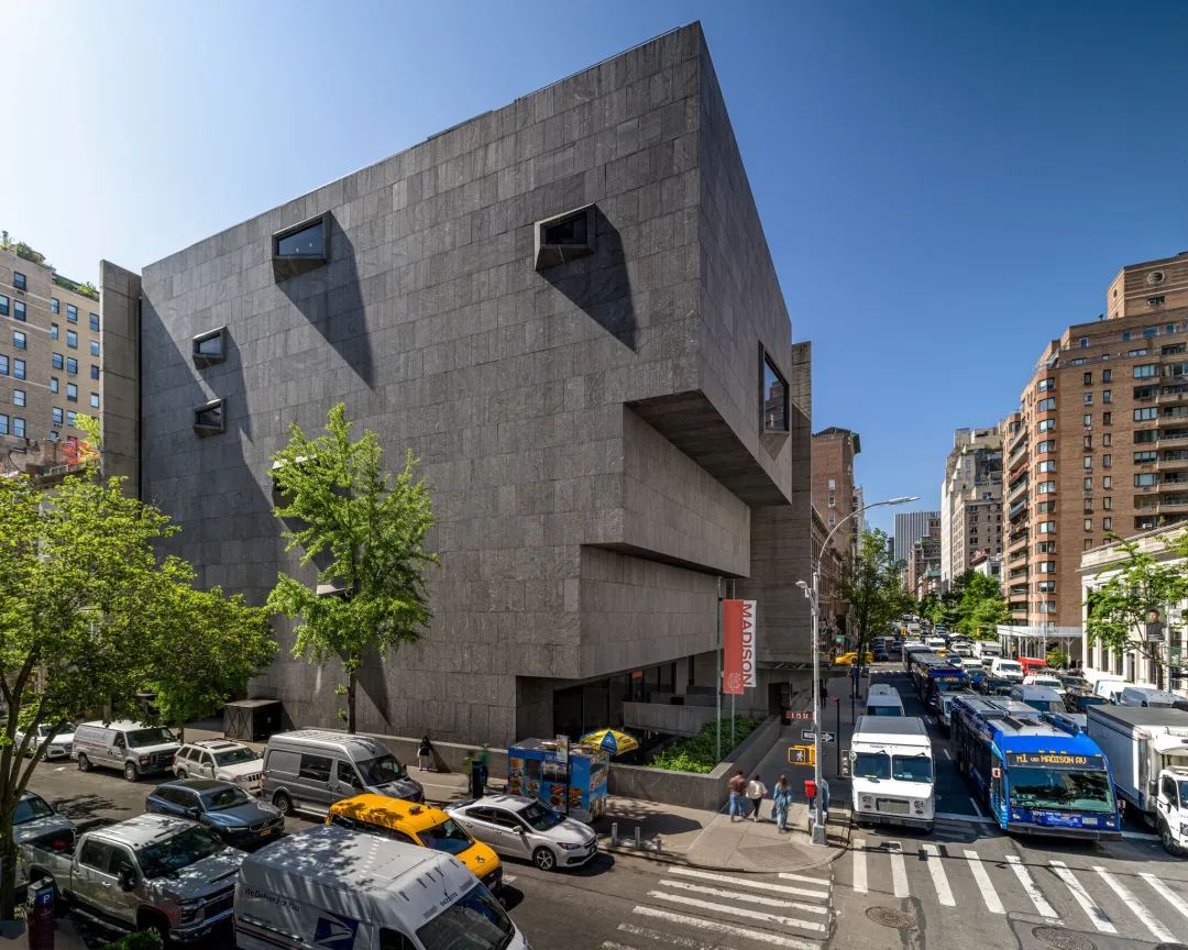 A busy street corner in New York City showing the exterior of a large stone building with geometric windows and shape.