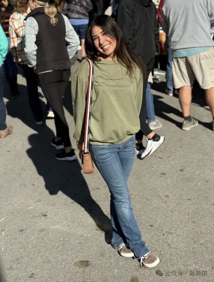 A young woman in a green sweatshirt and jeans stands outdoors.