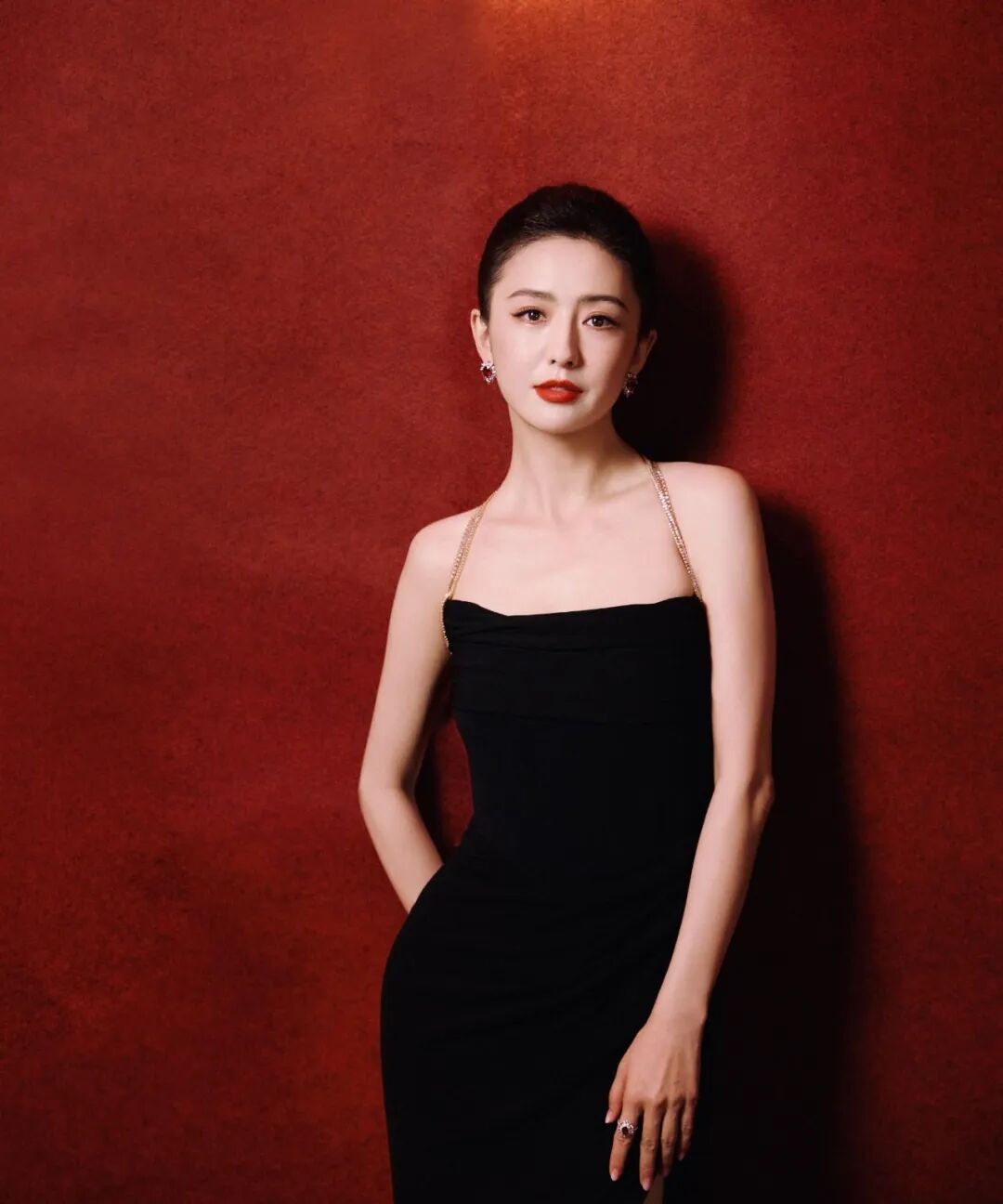 女性身着黑色侧开衩礼服，红唇妆容，背景红墙
