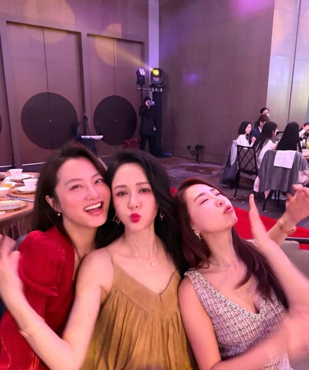 Three women pose together at a table, with one woman on the left smiling widely, the woman in the middle pouting her lips, and the woman on the right making a playful face