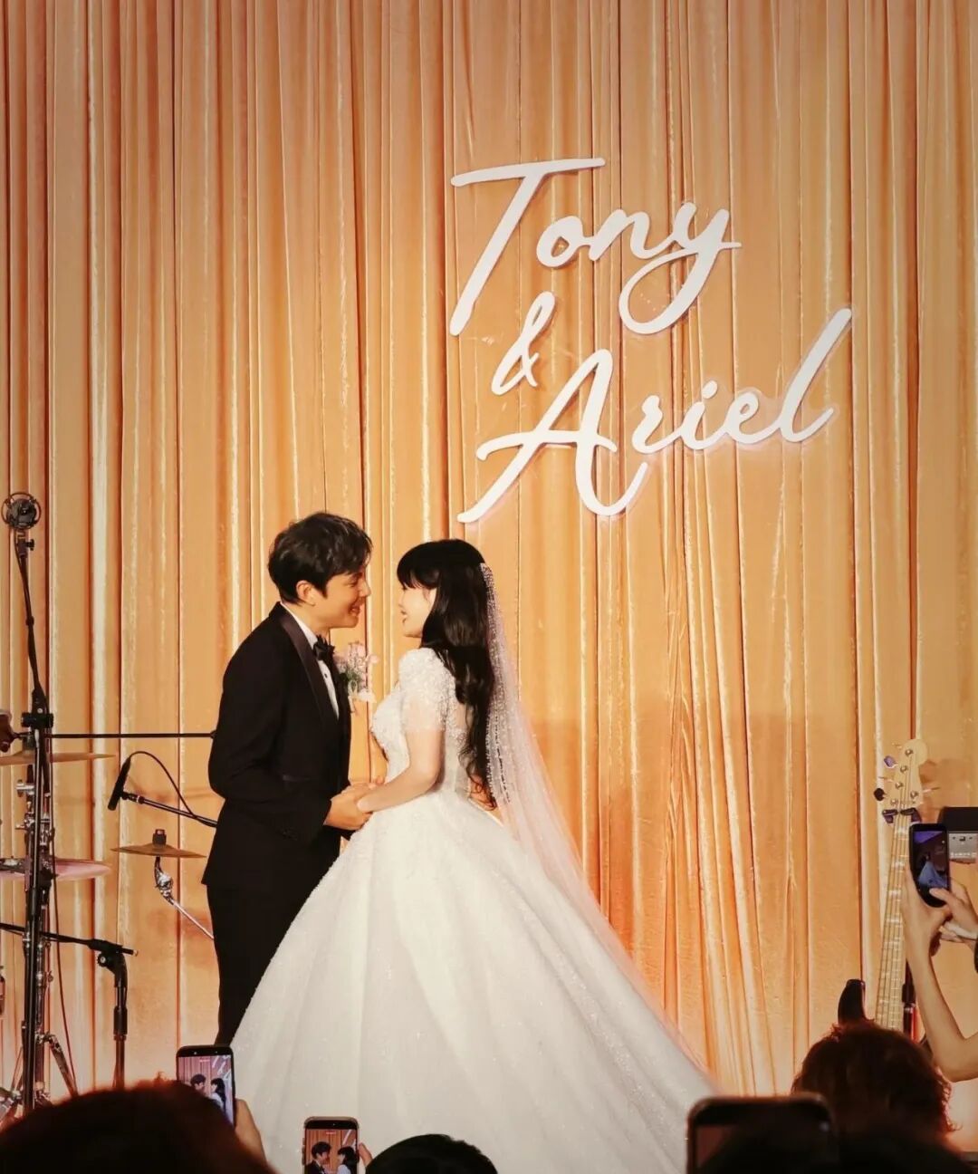 A bride and groom gaze at each other in front of a gold curtain and the words Tony & Ariel