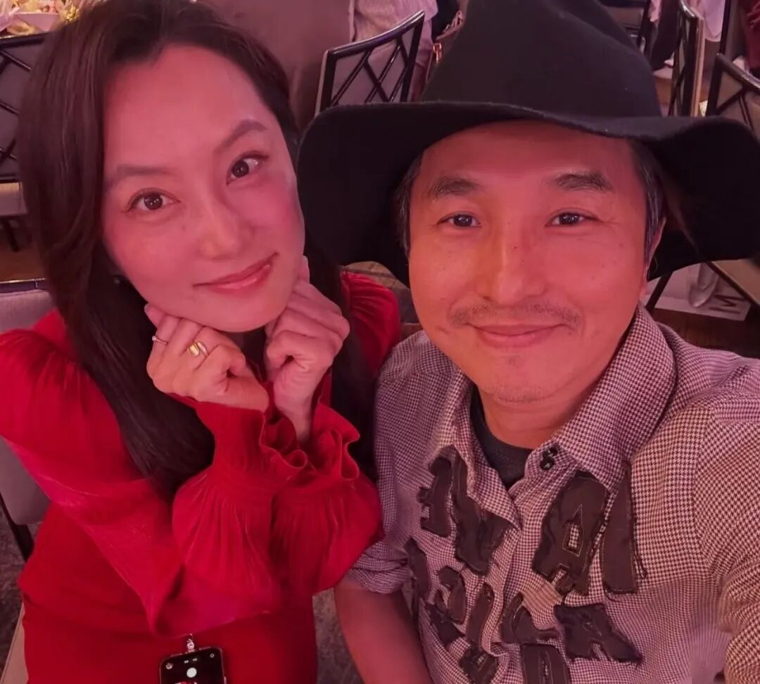 Two people, woman with hands under chin in red dress, and man with a black hat, are taking a selfie