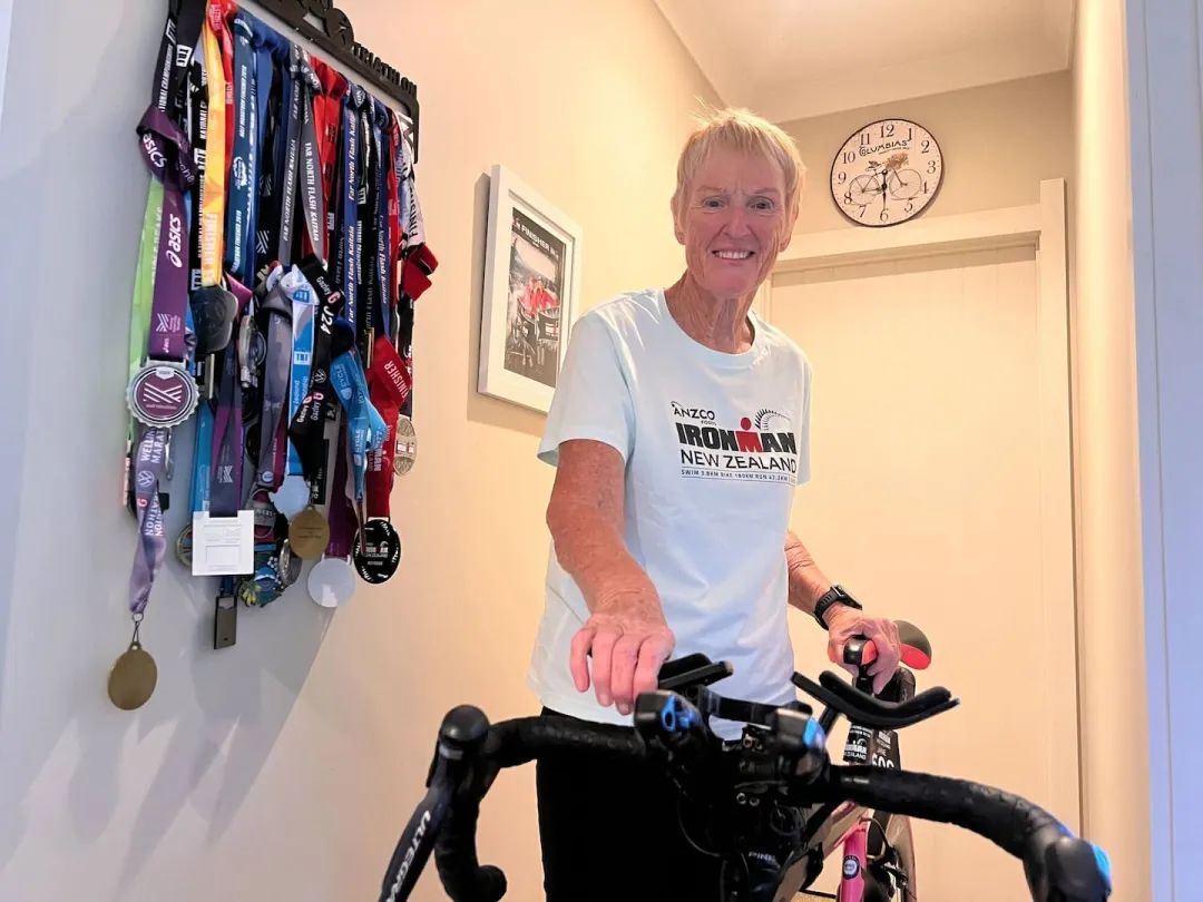 Jane Lee stands proudly by a wall of medals, reflecting on decades of competition, while a photo with her daughter Sarah Lee from a past Ironman reminds her that this journey has always been about family. Photo / Rafaella Melo