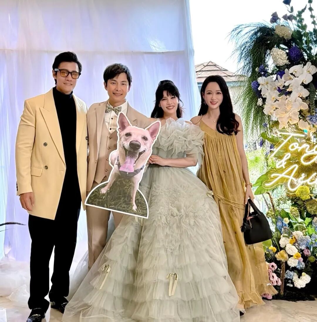 Group of people smiling in front of a floral archway and white curtain, including the bride in a green dress, a cardboard cutout of a dog, and the name 'Tony & Avi' in neon lights