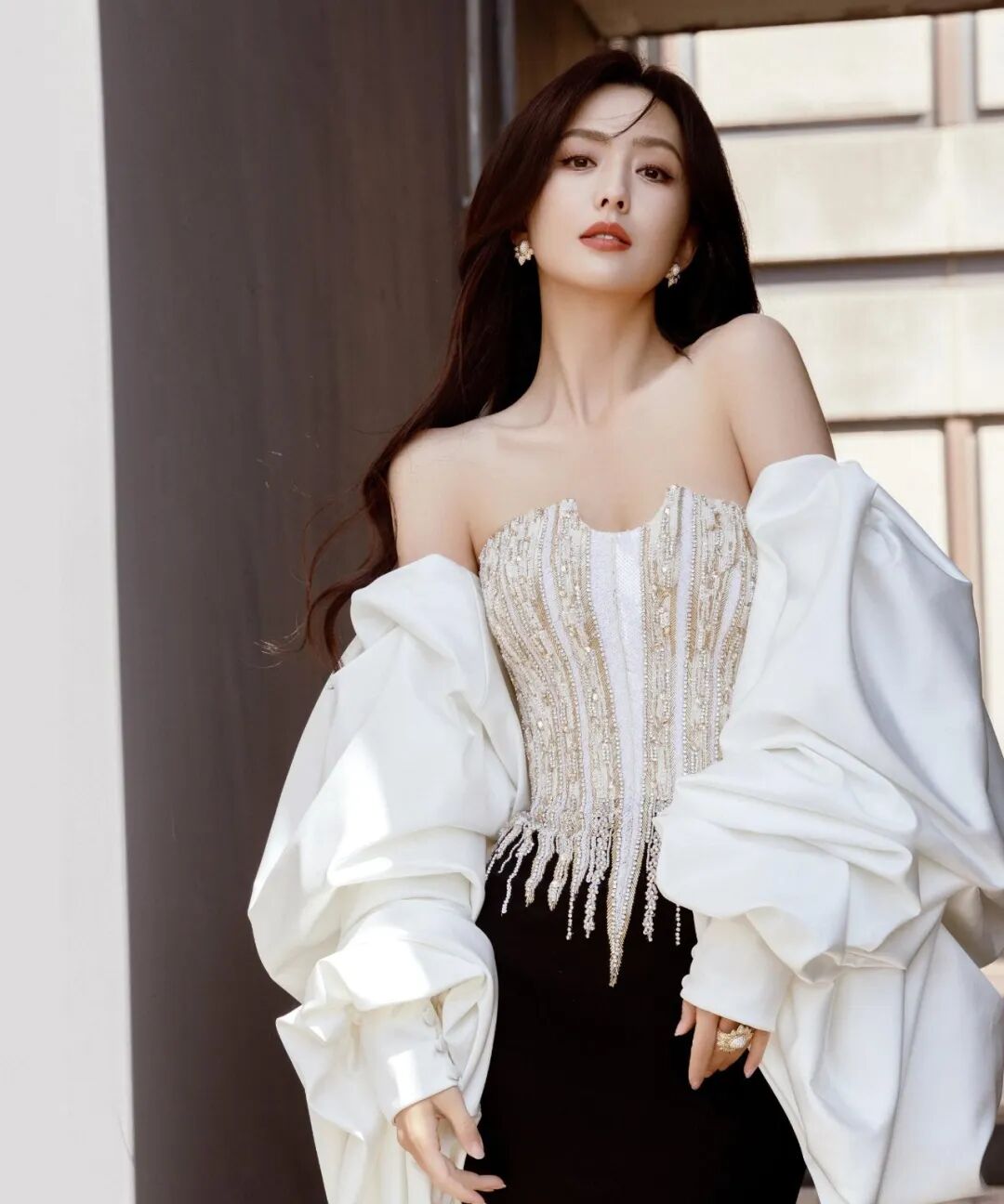 女性身着白金色礼服，白色蓬松袖子，在建筑前摆姿势