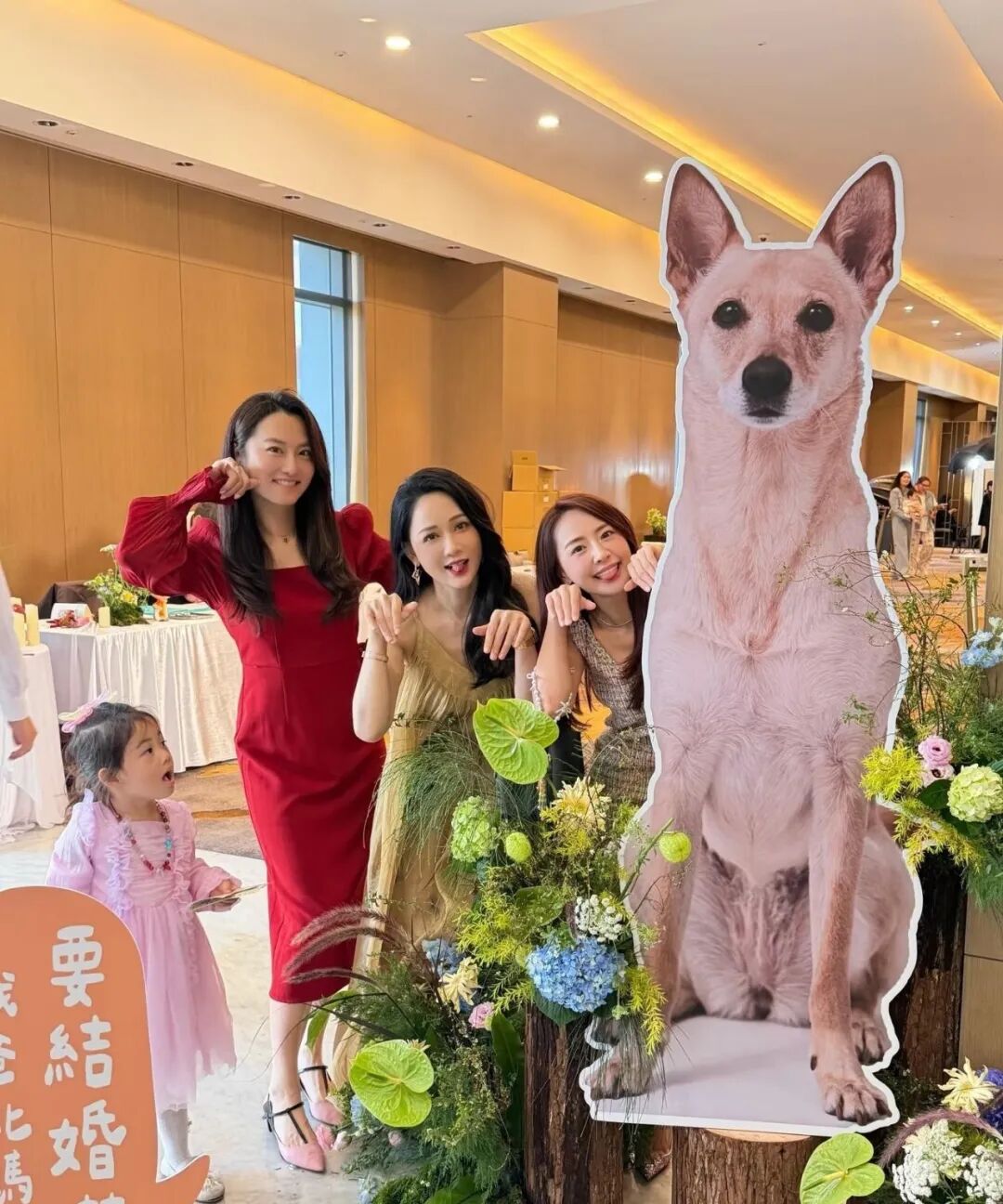 Three smiling women pose with a cardboard cutout of a dog at a wedding reception, while a young girl in a pink dress looks on