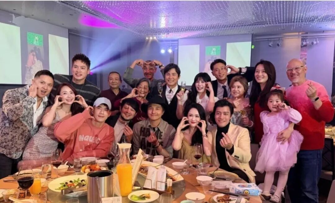 Group of people, mostly adults, gathering around a circular table during a celebration, making heart-shaped gestures with their hands