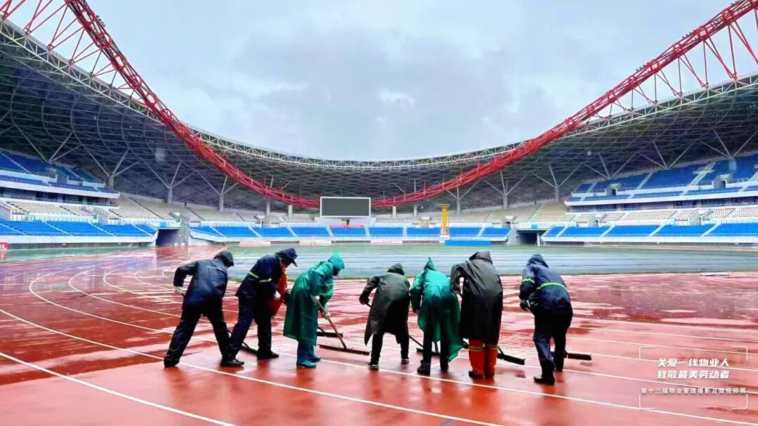 23.167.大庆府民物业管理雨中坚守，赛事护航 暴风雨突袭，体育场跑道很快积满了水。物业工作人员迅速行动，他们披上雨衣，拿起工具，冲向跑道。雨水顺着帽檐灌进脖颈，浸透的衣衫紧紧贴在身上，汗水和雨水混成一股，顺着下巴滴在地上。可他们毫不在意，专注地挥动工具，奋力将积水推向排水口，只为保障赛事活动的顺利进行。随着后一汪积水缓缓退去，他们直起早已酸痛的腰背，脸上浮现出欣慰的笑容。没等喘口气，他们又扛起工具，默默走向下一个需要维护.jpg