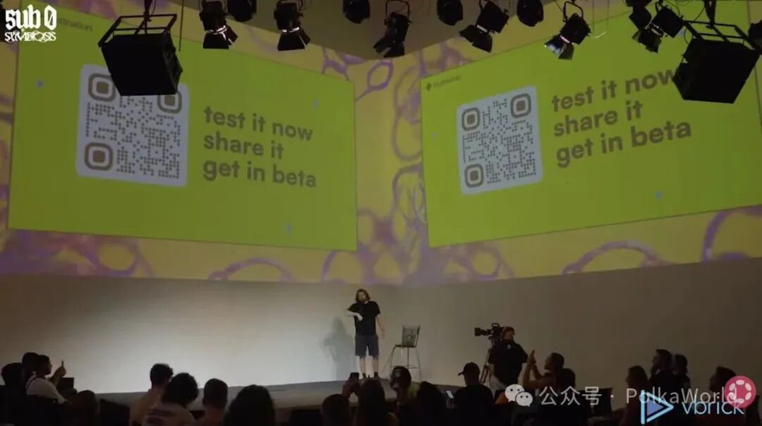 Conference stage with large yellow screens displaying QR codes and text reading test it now and get in beta surrounded by lighting rigs a person speaking at a podium in casual attire audience members seated and standing with cameras and vbrick logo visible on screens.