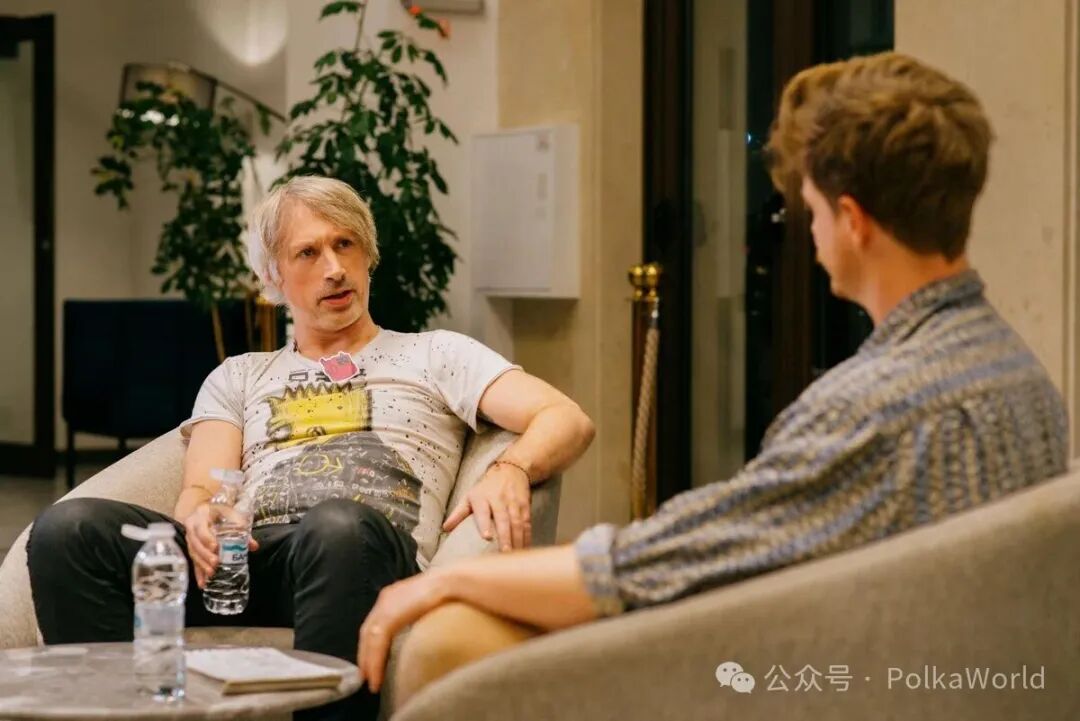 Two men sit facing each other in armchairs during a conversation in an indoor setting with plants and a door in the background. One man has light hair and wears a white t-shirt with a graphic print holding a water bottle. The other man has short brown hair and wears a light-colored patterned shirt. A table with papers is between them.