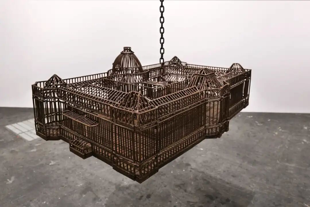 白色墙壁画廊空间中的雕塑装置细节，这些雕塑通过链条悬挂在天花板上，每个雕塑都仿照殖民地时期的机构建筑