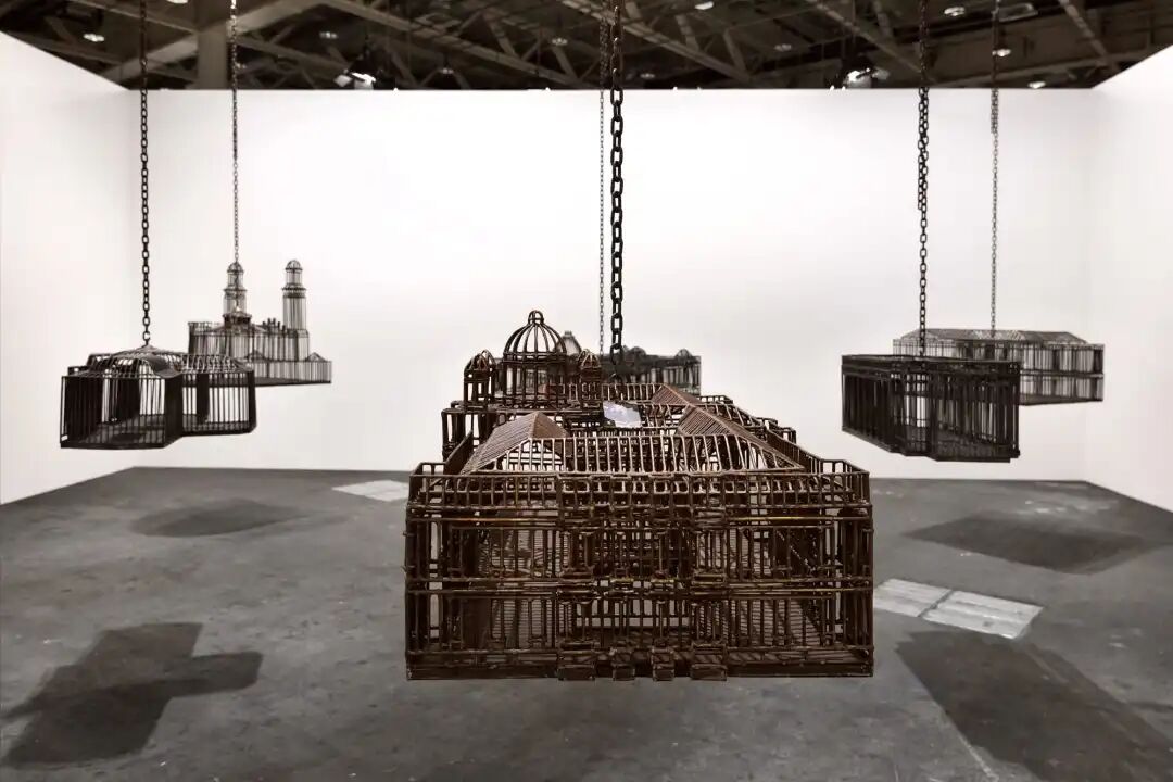 白色墙壁画廊空间中的雕塑装置细节，这些雕塑通过链条悬挂在天花板上，每个雕塑都仿照殖民地时期的机构建筑