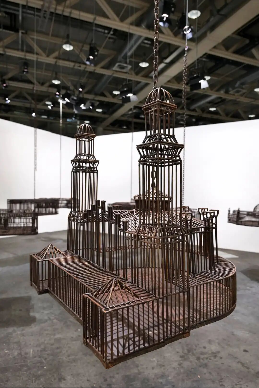 白色墙壁画廊空间中的雕塑装置细节，这些雕塑通过链条悬挂在天花板上，每个雕塑都仿照殖民地时期的机构建筑