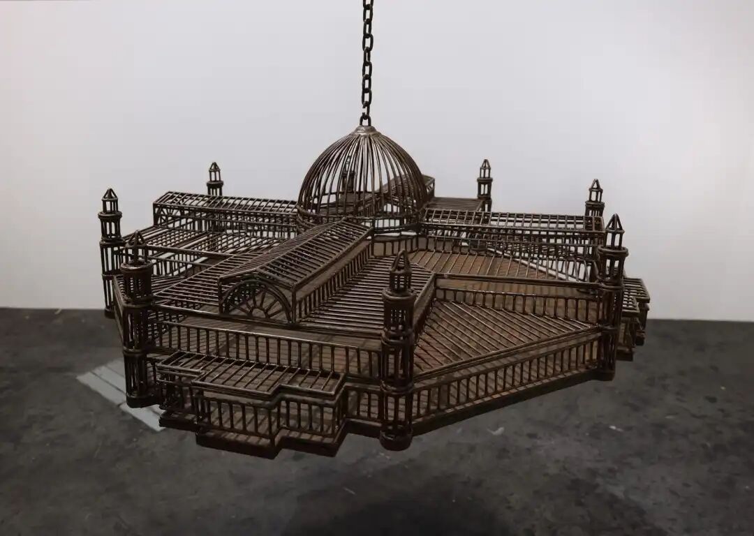 白色墙壁画廊空间中的雕塑装置细节，这些雕塑通过链条悬挂在天花板上，每个雕塑都仿照殖民地时期的机构建筑