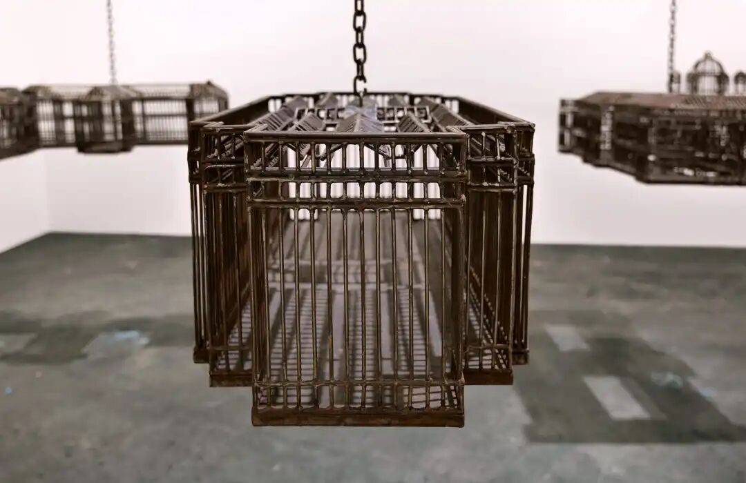 白色墙壁画廊空间中的雕塑装置细节，这些雕塑通过链条悬挂在天花板上，每个雕塑都仿照殖民地时期的机构建筑