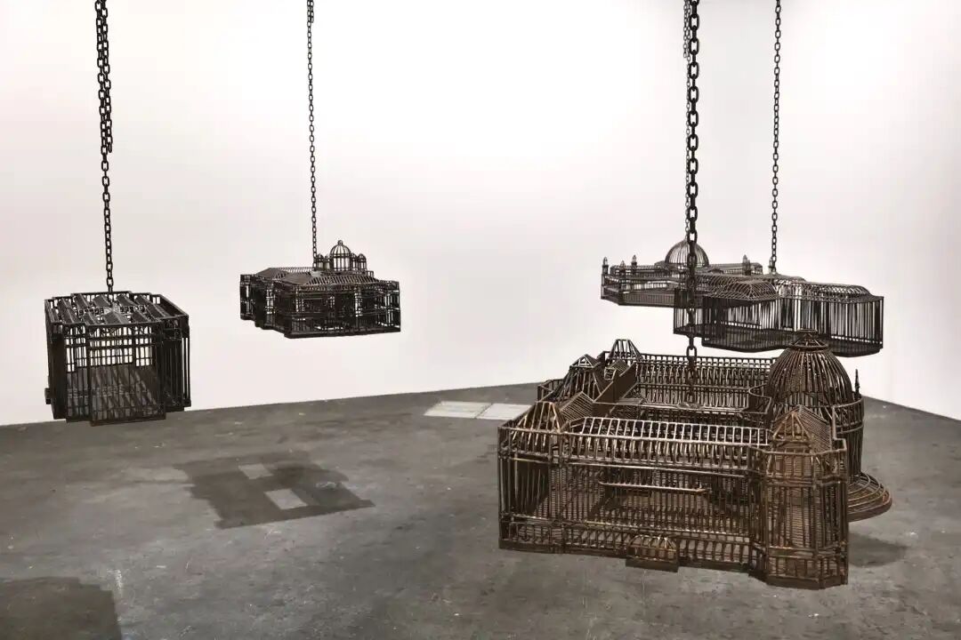 白色墙壁画廊空间中的雕塑装置细节，这些雕塑通过链条悬挂在天花板上，每个雕塑都仿照殖民地时期的机构建筑