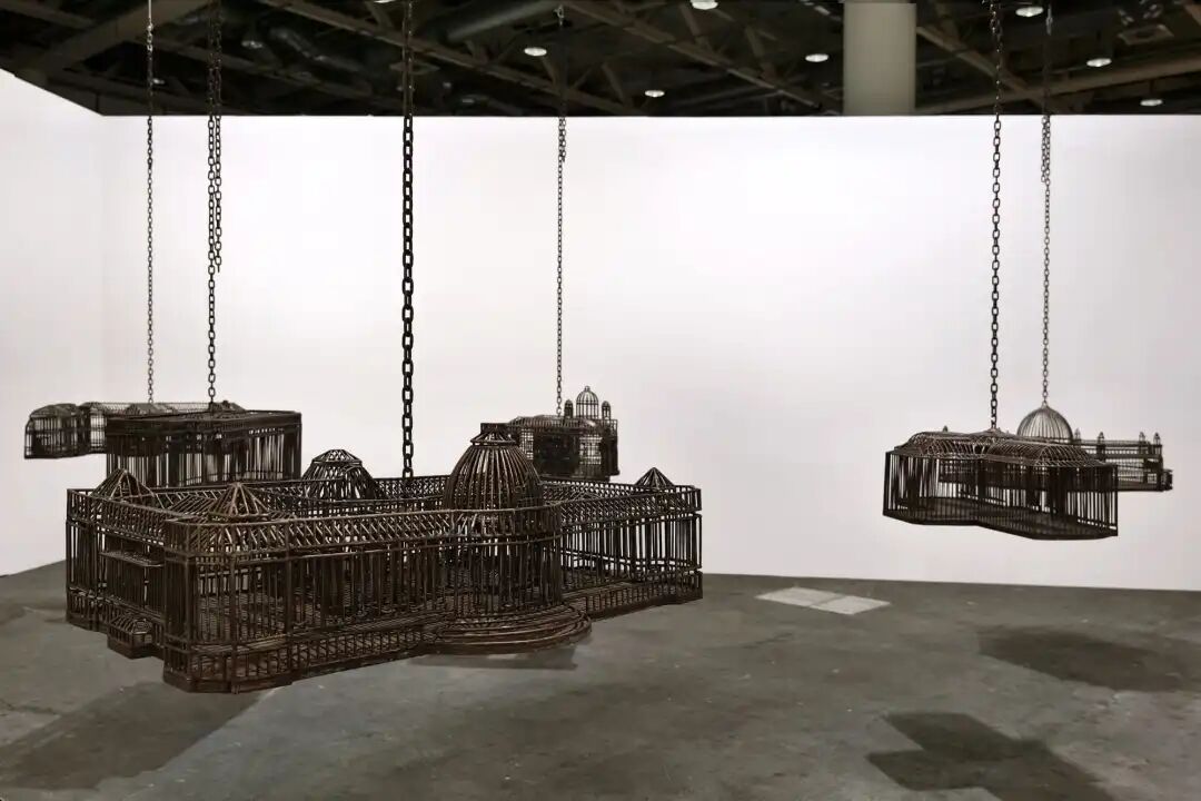 白色墙壁画廊空间中的雕塑装置细节，这些雕塑通过链条悬挂在天花板上，每个雕塑都仿照殖民地时期的机构建筑