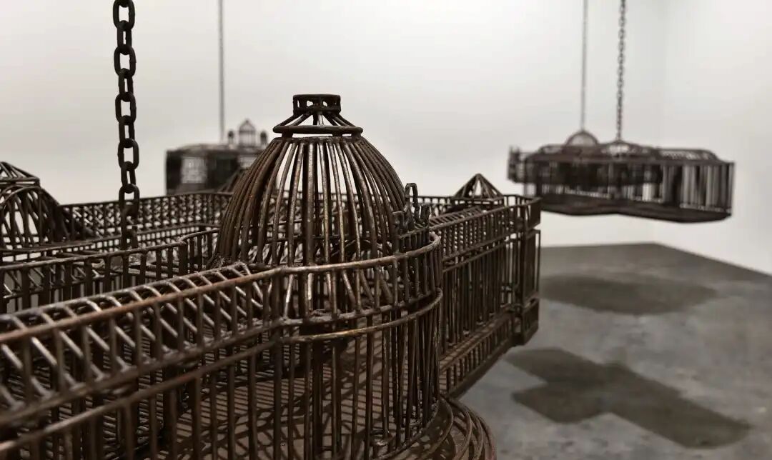 白色墙壁画廊空间中的雕塑装置细节，这些雕塑通过链条悬挂在天花板上，每个雕塑都仿照殖民地时期的机构建筑