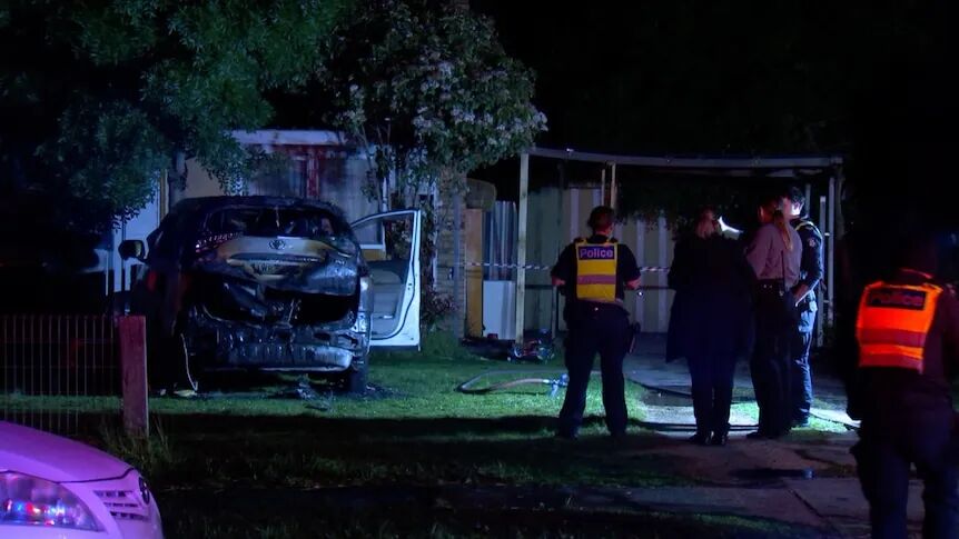 Police officers stand in a driveway looking at a burnt out car at night.
