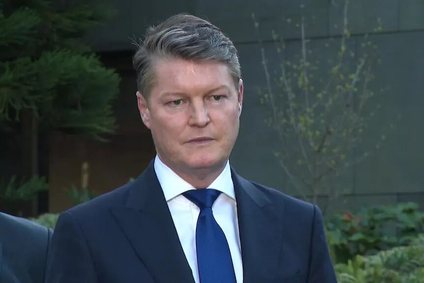 A man in a navy blazer, white collared shirt and blue tie stares just off camera.
