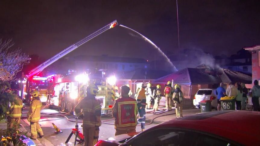 The scene of a house fire shows firefighters standing under bright lights at night