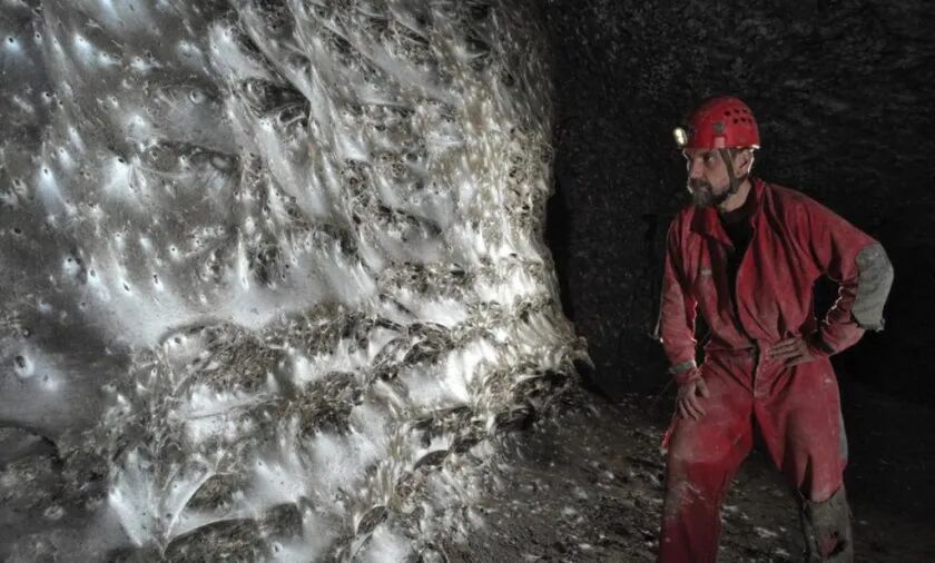 A man in a red jumpsuit examines a giant spider web in a dimly lit cave.