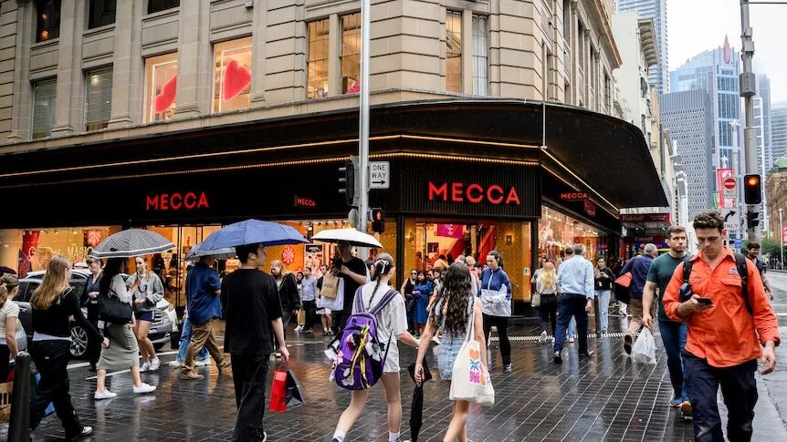 A crowd of people carrying umbrellas and shopping bags walk along a street on a rainy day.