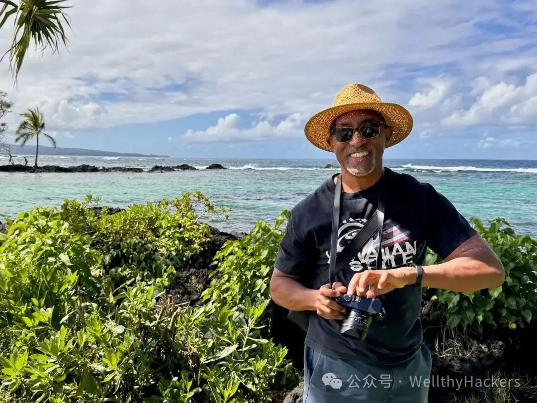 Michael in sunglasses and a straw hat holds a camera while standing on a coastal trail at Carlsmith Beach Park in Hawaii. Behind him, turquoise ocean water and palm trees reflect a sunny, relaxed atmosphere.