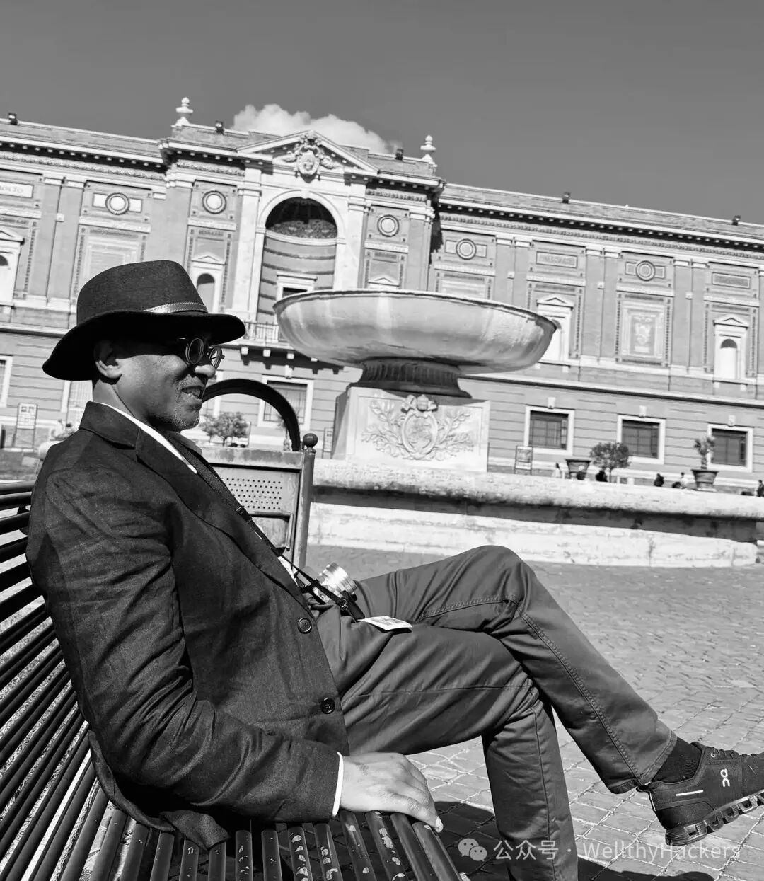 A man wearing a dark blazer, hat, and sunglasses relaxes on a bench outside the Vatican Museums, with a large fountain and ornate Renaissance-style building in the background. He appears calm and reflective in the midday sun.