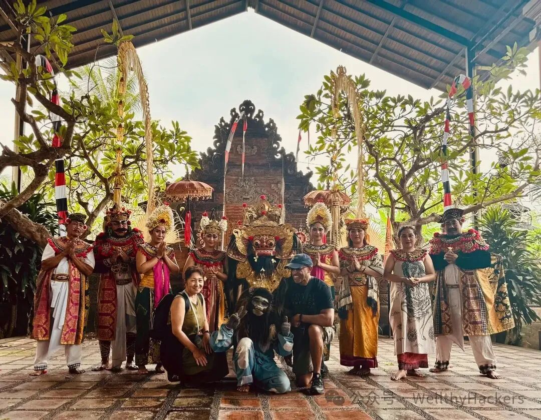 A man and woman kneel in front of a group of Balinese performers dressed in traditional ceremonial costumes, framed by tropical trees and a temple structure. The setting is vibrant and cultural, evoking travel, tradition, and celebration.