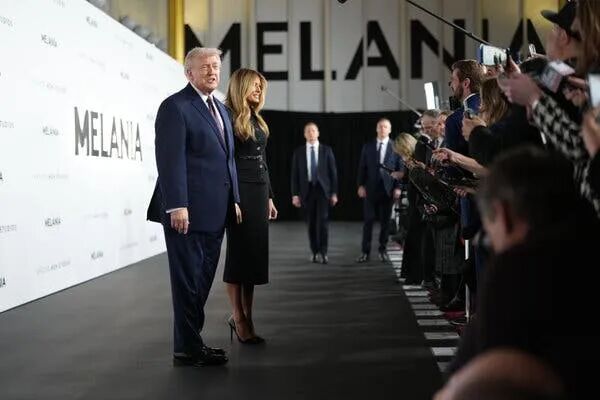 President Trump, wearing a navy suit, poses beside Melania Trump, in a black suit with a skinny belt, in front of gathered journalists.