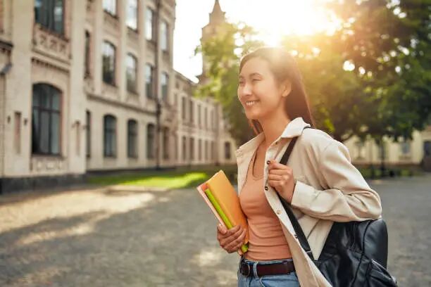 A young woman student walking on campus with a backpack and books, enjoying the sunshine A girl embodies hope and dedication to learning as she walks through the university campus in the afternoon, carrying books and a backpack under the sunshine, symbolizing academic aspirations international students stock pictures, royalty-free photos & images