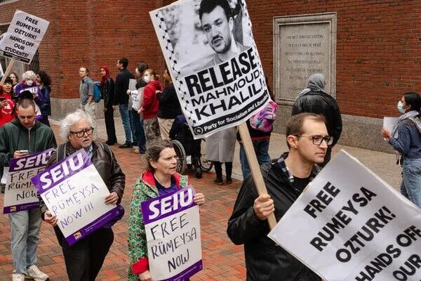 People walk in a picket line holding signs protesting the targeting of international students.
