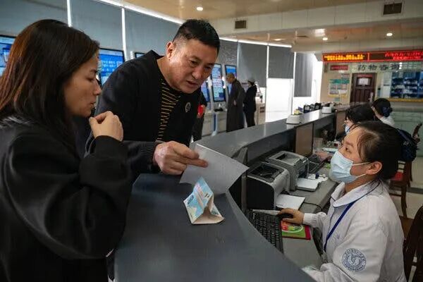 A man shows papers to a seated individual in a white coat and face mask at a counter.  A woman stands next to them, looking on.