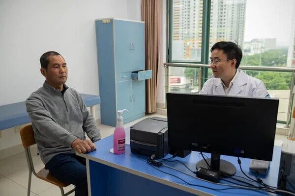 Two people sit at a blue desk in a room. One in a grey sweater; the other in a white lab coat with glasses.