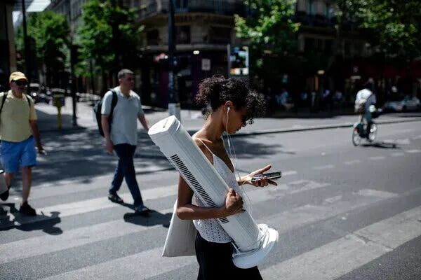 A person crosses a street, carrying a large white tower fan. They look at their smartphone; other people are in the background.