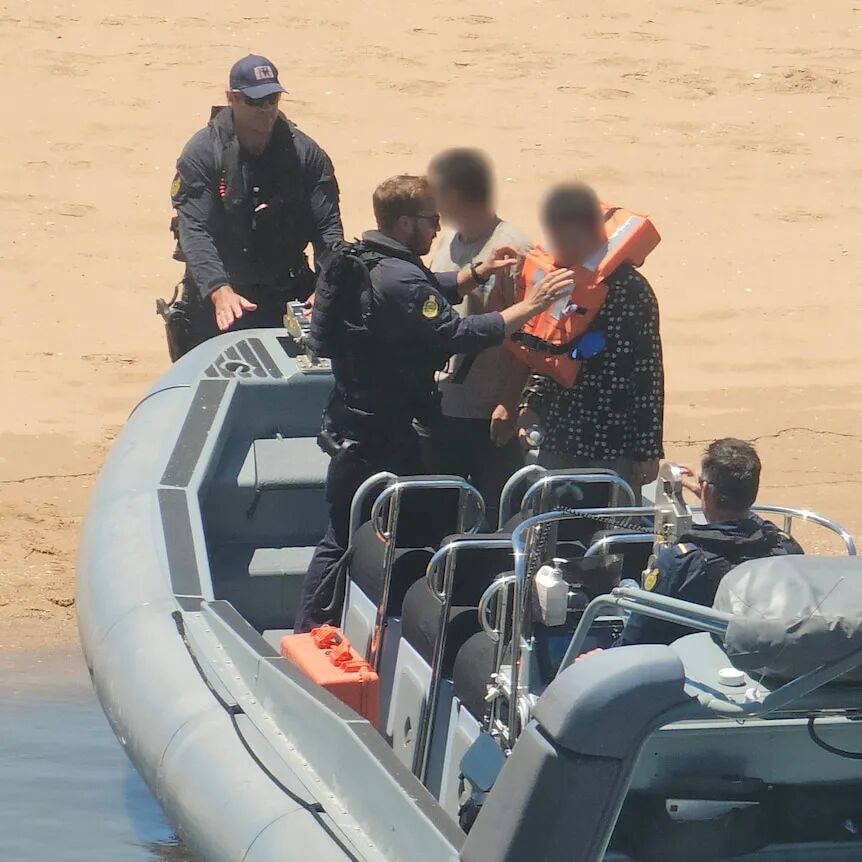 Men in uniiform hand lifejackets to other men on a boat moored on orange sand.