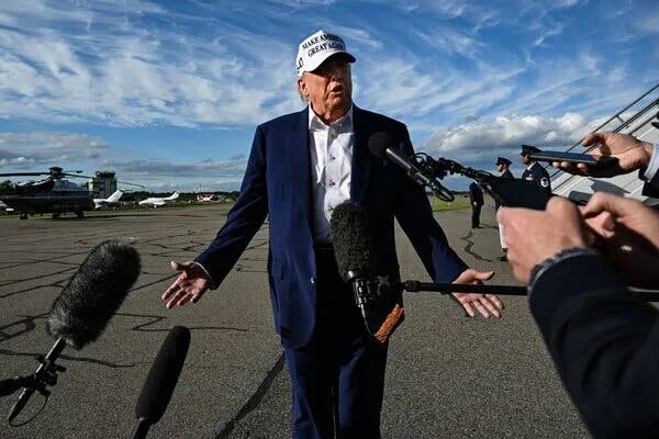 President Trump, wearing a white Make America Great Again cap, spreads his arms while speaking on a tarmac. A series of microphones are thrust toward him.