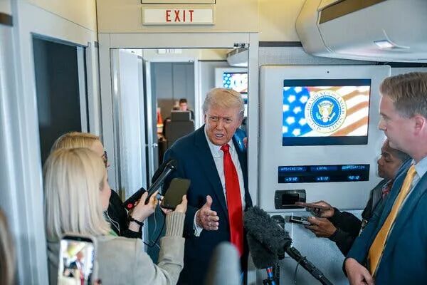 President Trump leaning in a doorway on a plane and talking as journalists hold microphones in front of him.