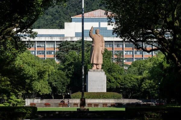 A statue of Mao Zedong stands amid large leafy trees. Behind it is a campus building with an orange peaked roof.