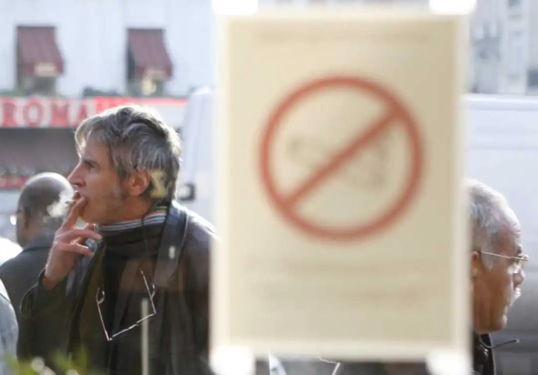 A man smokes a cigarette outside a brasserie, in front of a sticker on a window reading: "Smoking is forbidden,"
